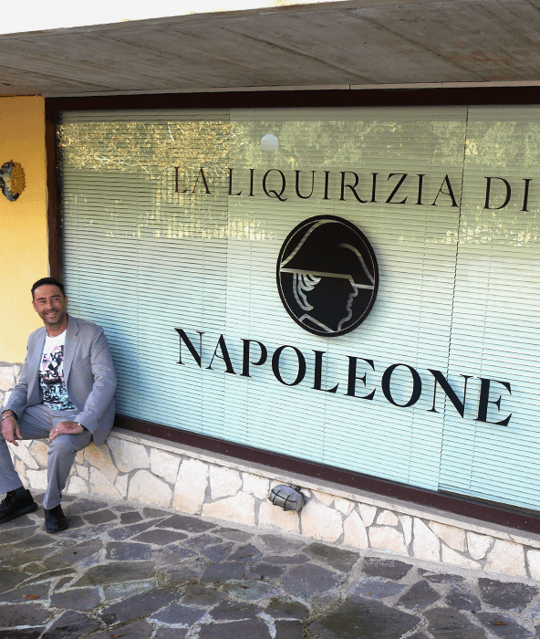 Ragazzo sorridente con t-shirt bianca e stampa fantasia con giacca grigia seduto su uno scalino davanti all'insegna a vetro con il logo de La Liquirizia di Napoleone